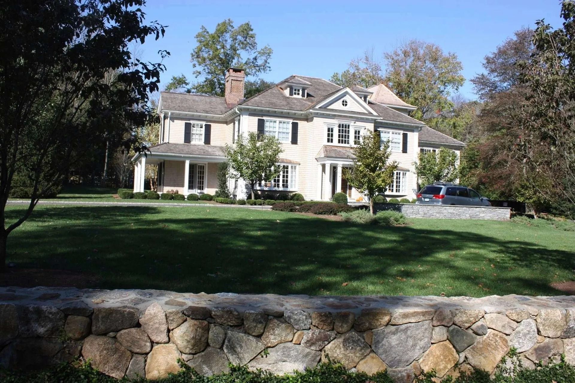 Large white house with a front lawn, stone wall, and trees under a clear sky