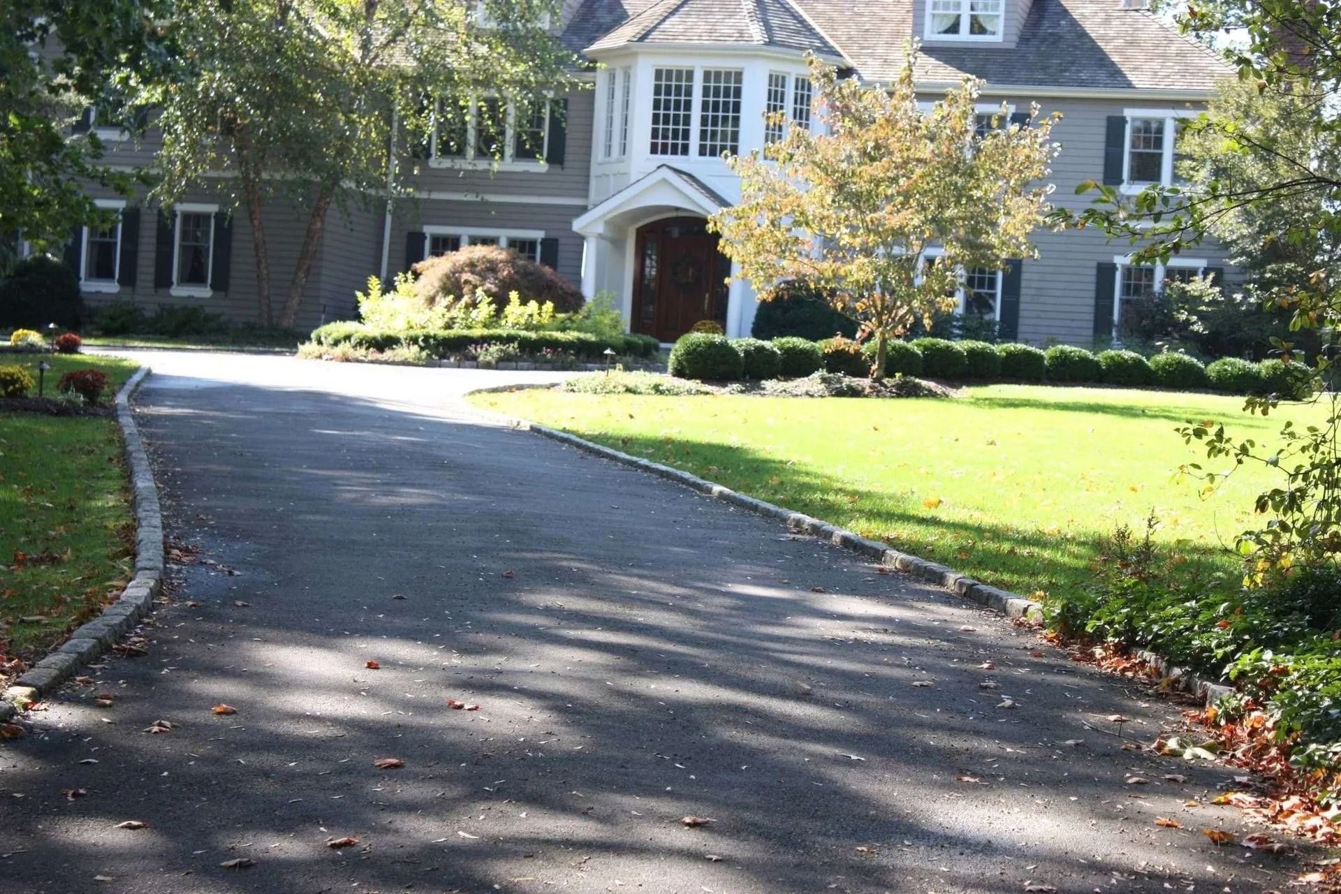 Curved driveway leading to a gray house with white columns and a green lawn in sunlight