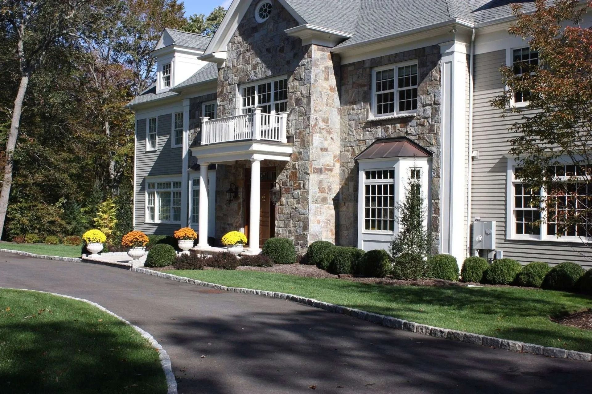 Large gray stone house with white trim, front porch, yellow chairs, and a curved driveway lined by trees