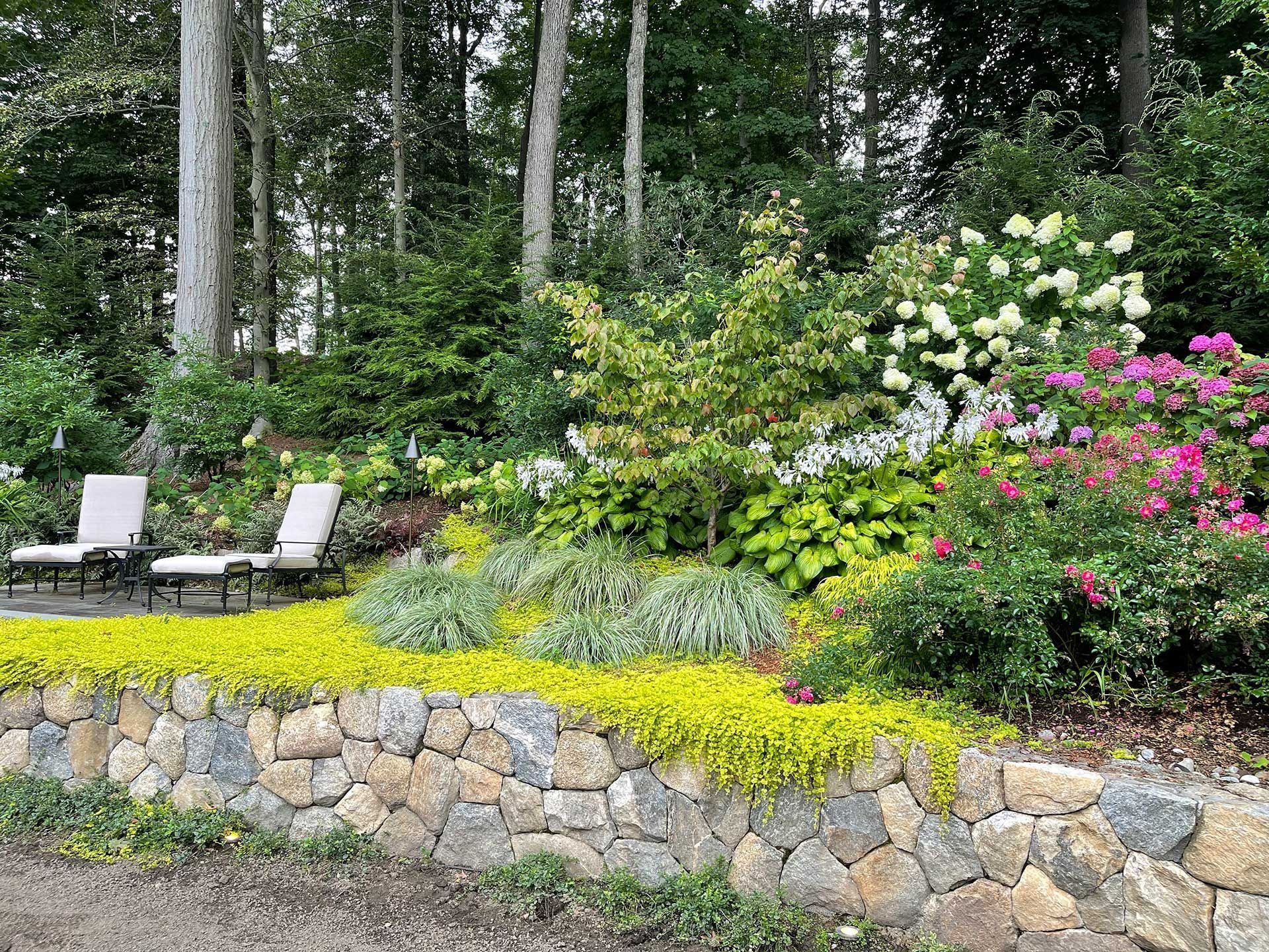 Stone retaining wall below a lush garden with white lounge chairs, pink flowers, and tall trees behind.
