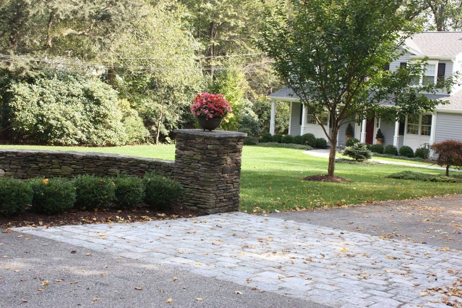 Driveway and landscaped front yard with stone pillar, red flowers, trees, and a white house in the background