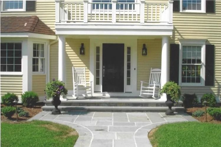 Front porch of a yellow house with white trim, steps, potted plants, and two white rocking chairs