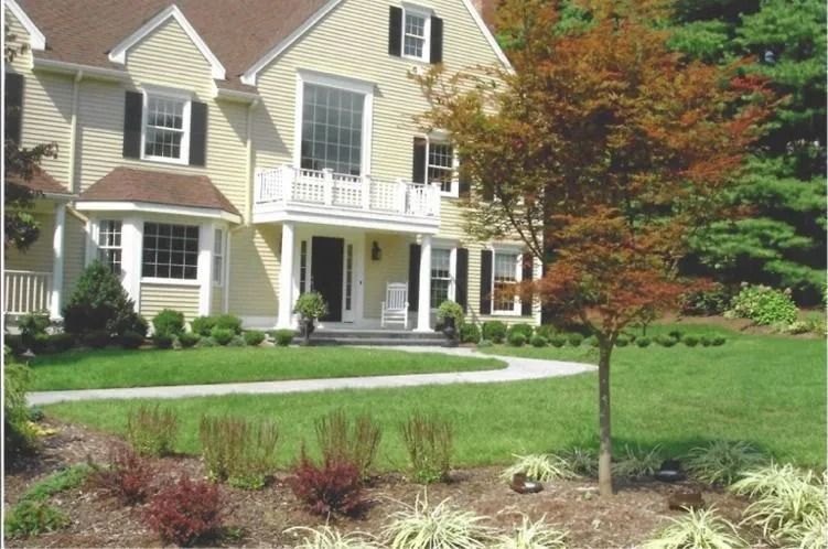 Suburban house with beige siding, white porch, manicured lawn, and autumn-colored tree in front yard