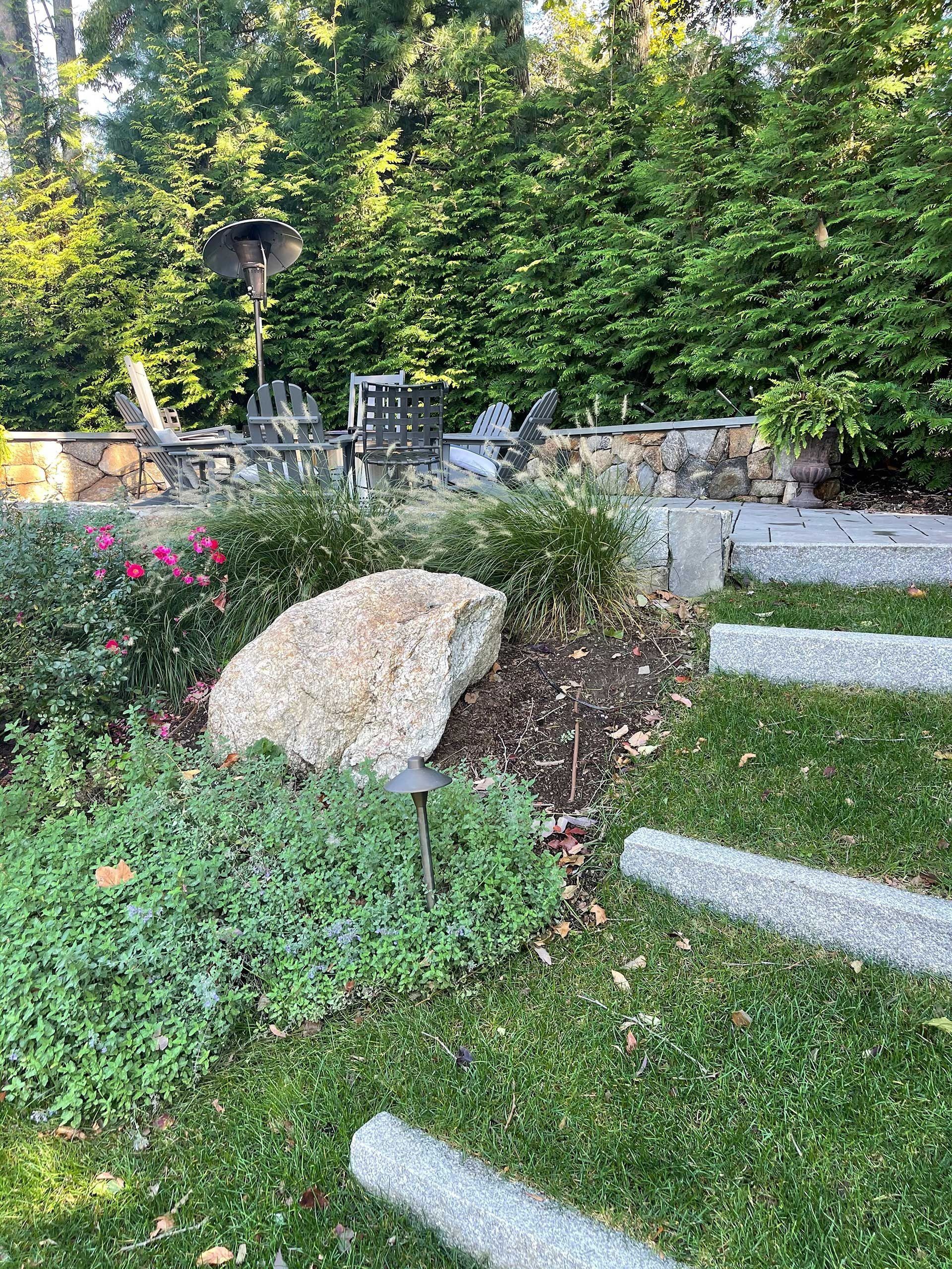 Stone steps in a green garden beside a large rock, flowers, and dense trees.
