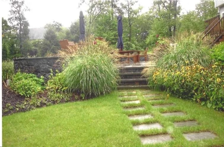 Rainy garden with stone steps, stepping stones, lush green lawn, and tall grasses beside a patio umbrella