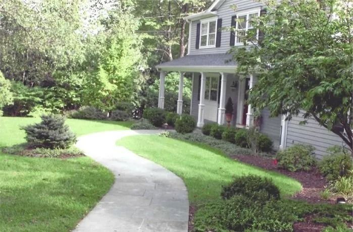 Curved concrete walkway leading to a gray house with a front porch, surrounded by lush green landscaping