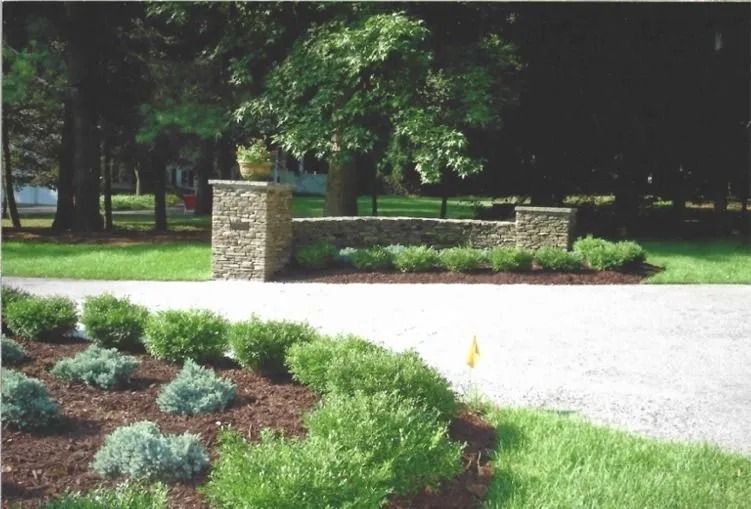 Landscaped park entrance with stone pillars, brick wall, and green shrubs beside a paved path