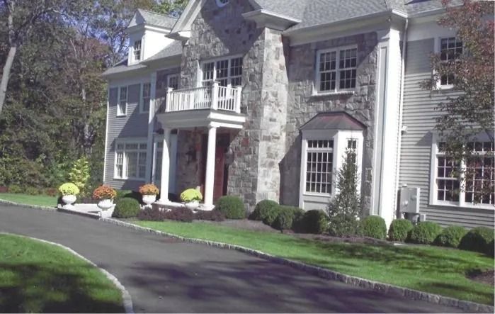 Large suburban house with stone columns, white siding, and a curved driveway bordered by manicured shrubs.