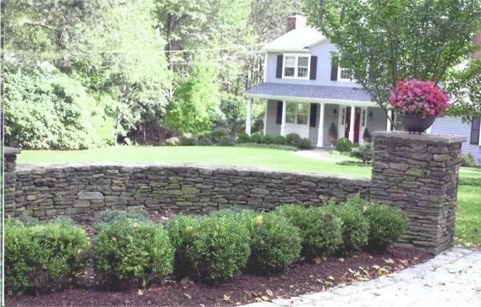 Stone retaining wall and landscaped yard in front of a blue house with white trim and porch.