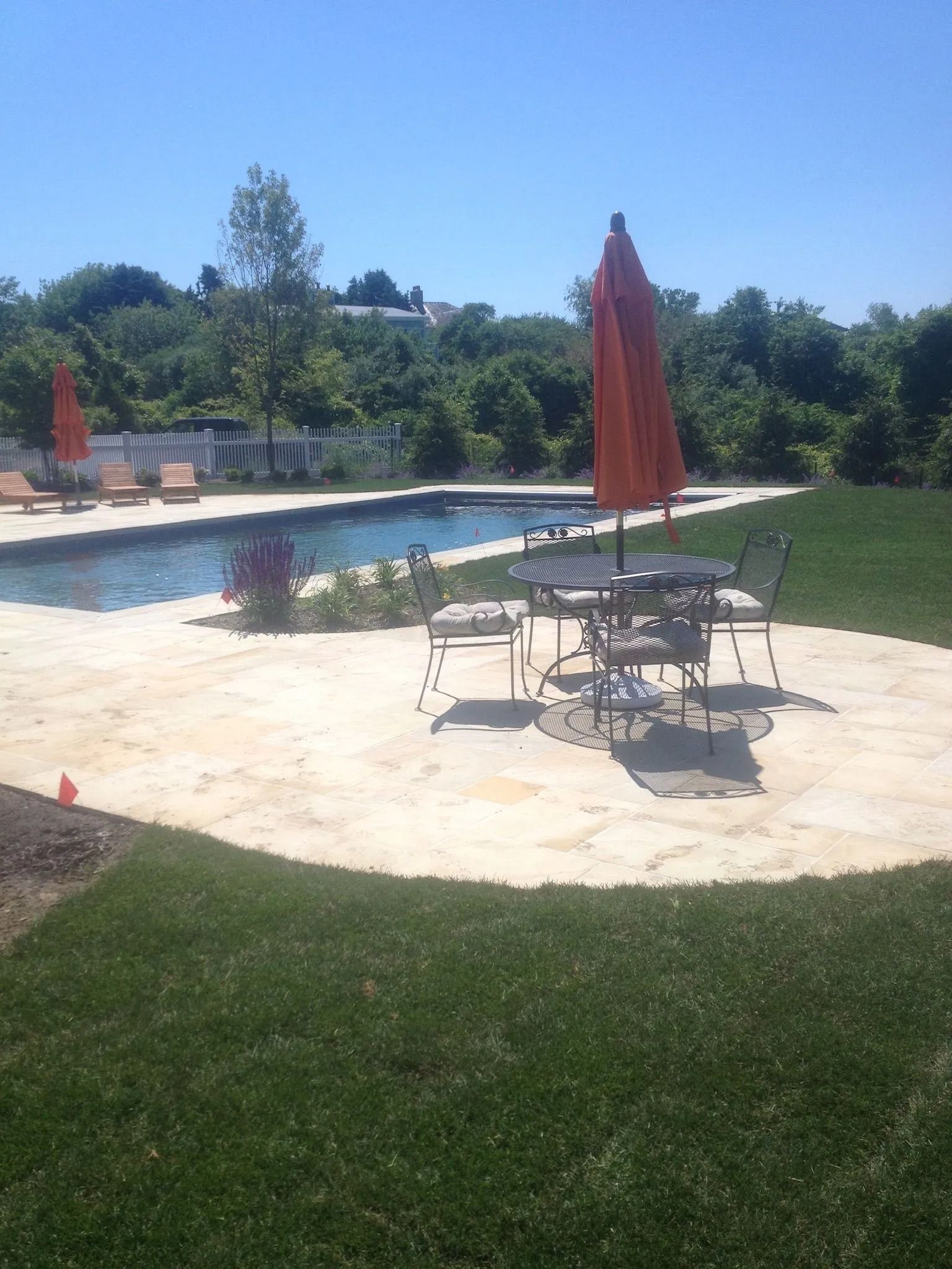 Sunny poolside patio with red umbrellas, tables, and chairs beside a grassy lawn and blue swimming pool