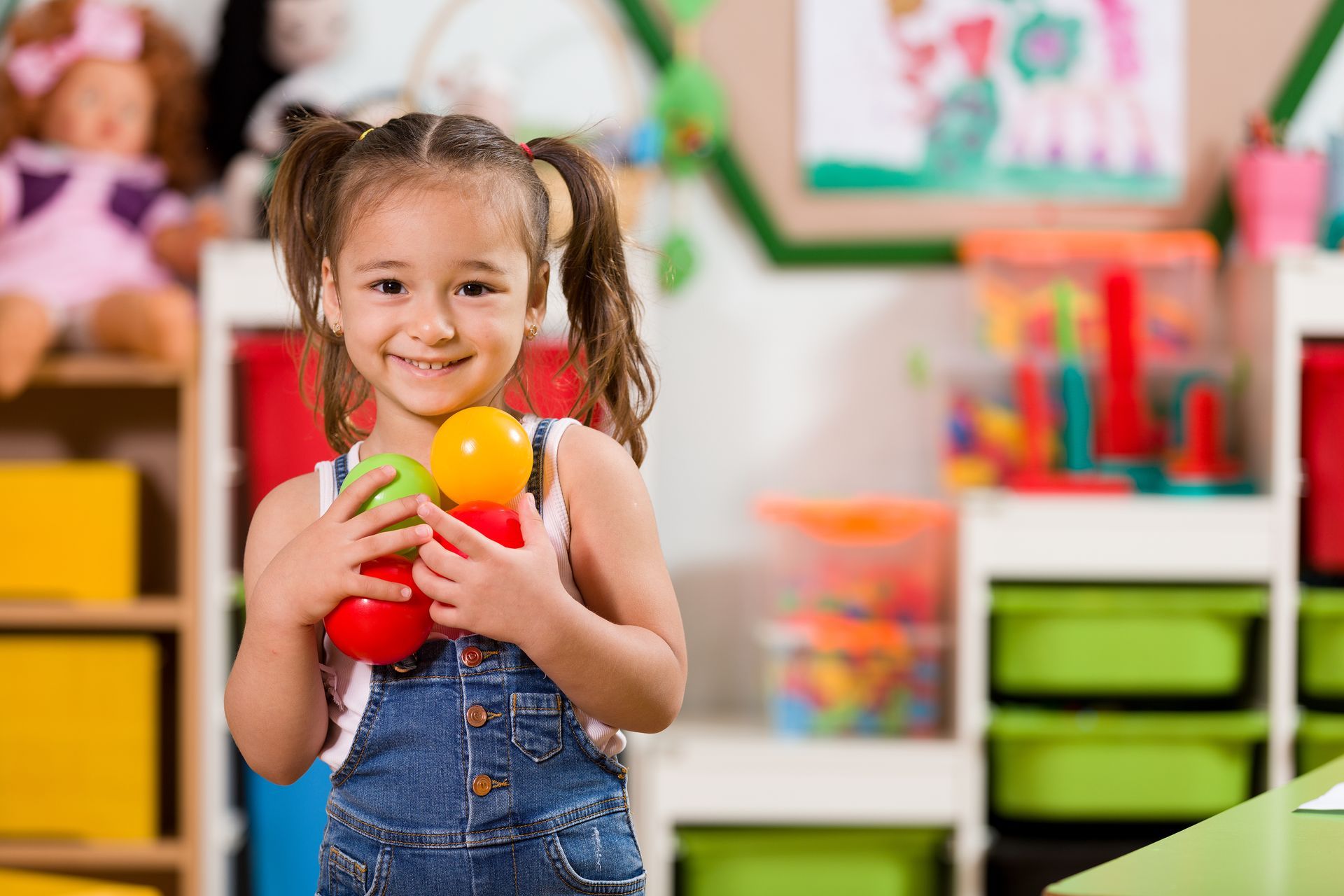 A young girl in a classroom smiles while holding colorful toys in her hands.
