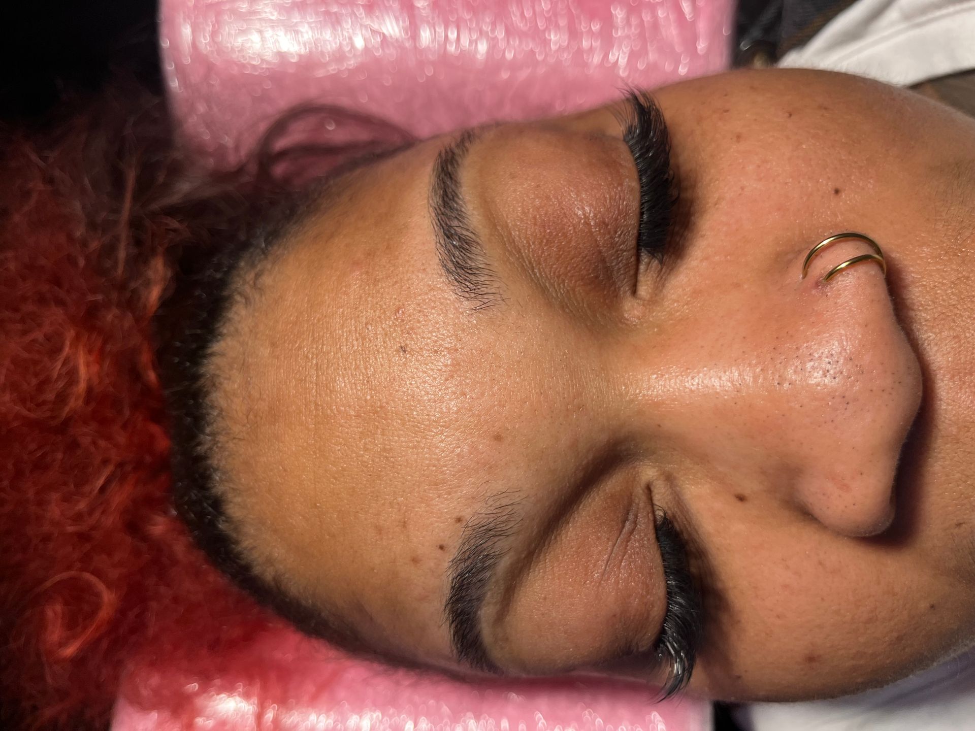Person with closed eyes, nose ring, dark eyebrows, resting on pink surface.
