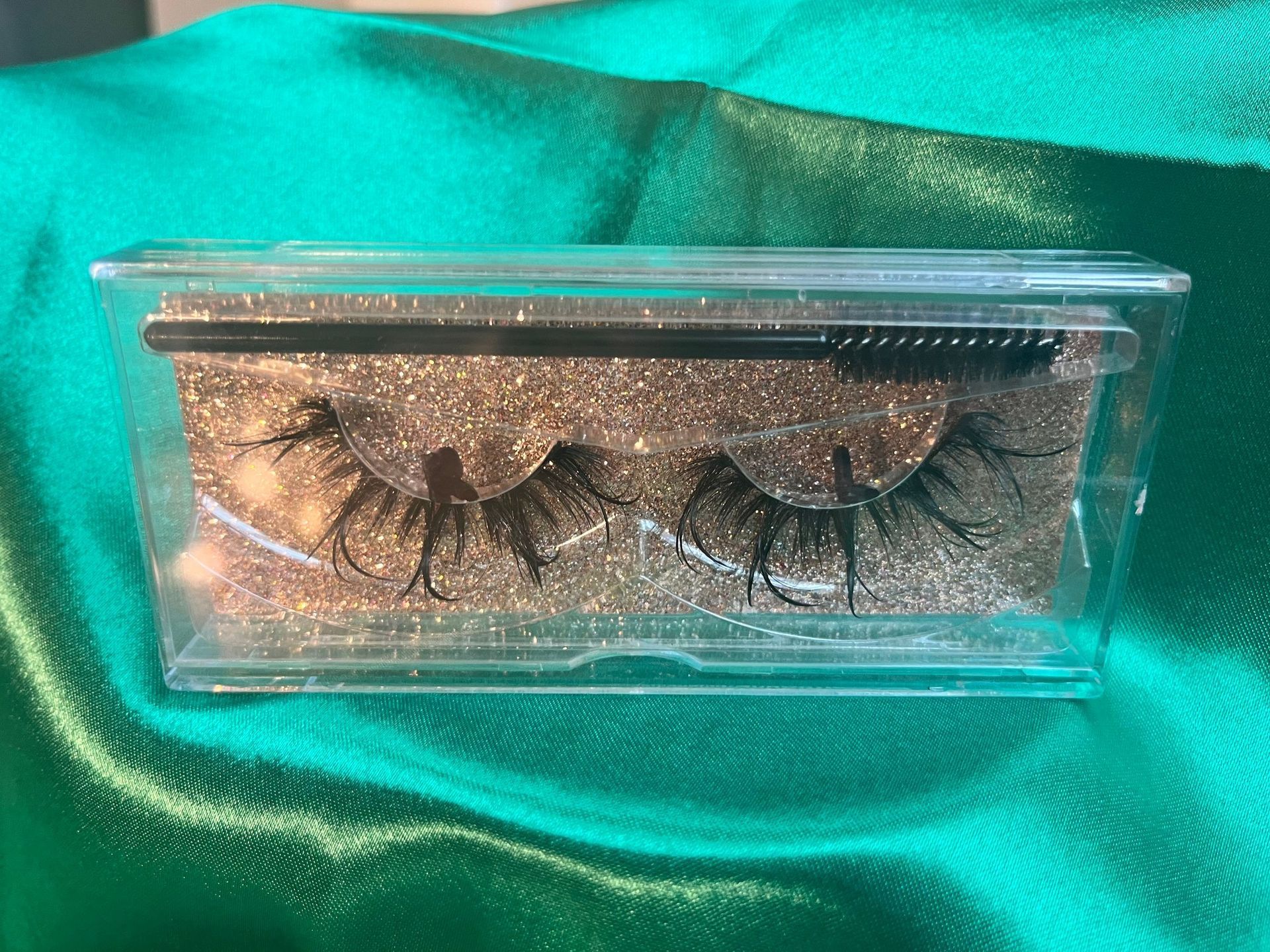 Clear box of false eyelashes on gold glitter background with brush, sitting on green satin.
