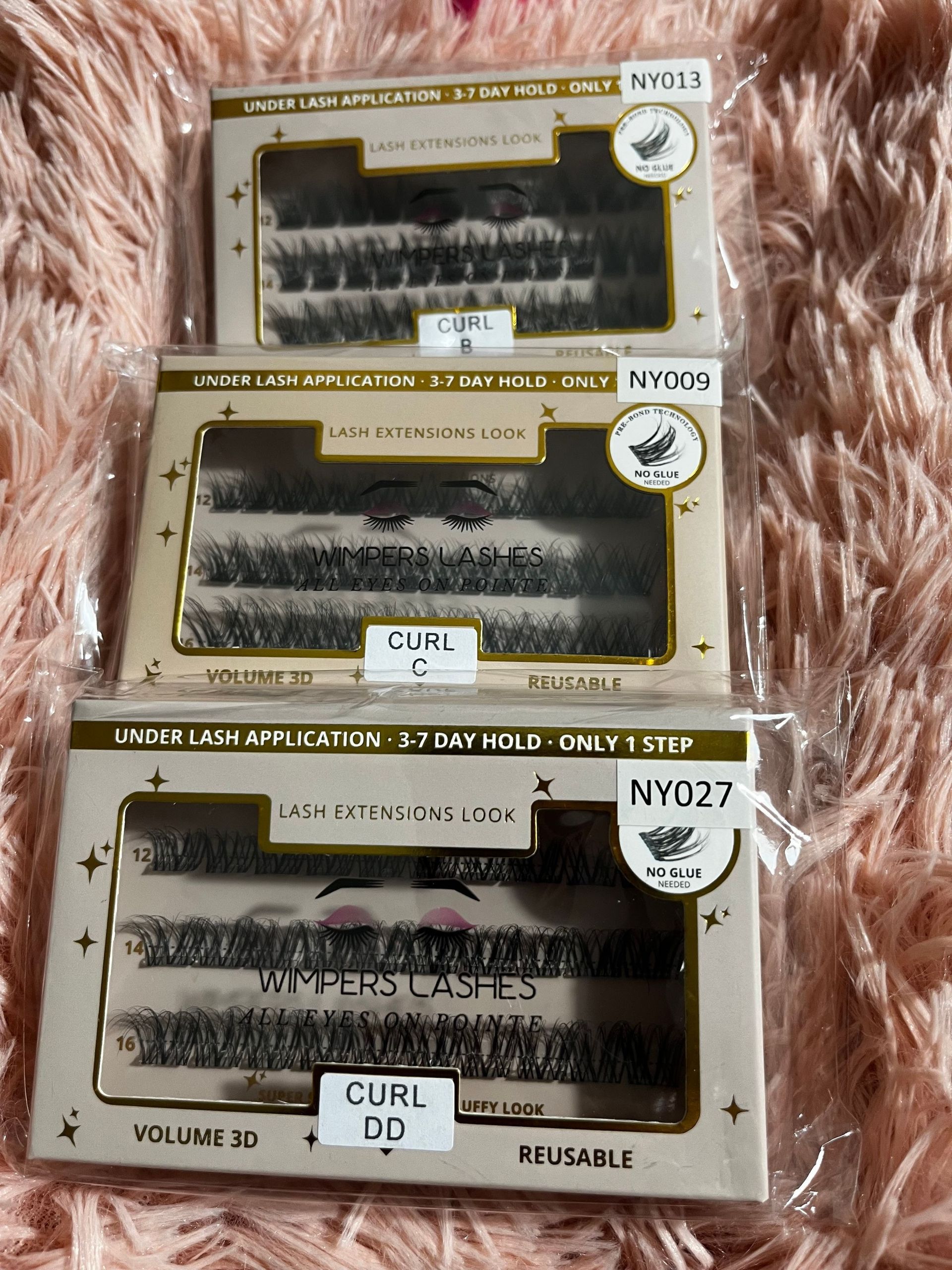Three boxes of false eyelashes on a pink surface. Each box is labeled with a "curl" type and style.