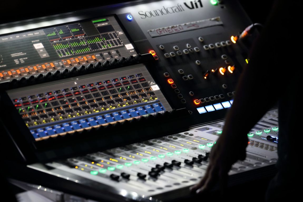 Soundboard with illuminated controls; a person's hand reaches for faders, dimly lit stage setting.