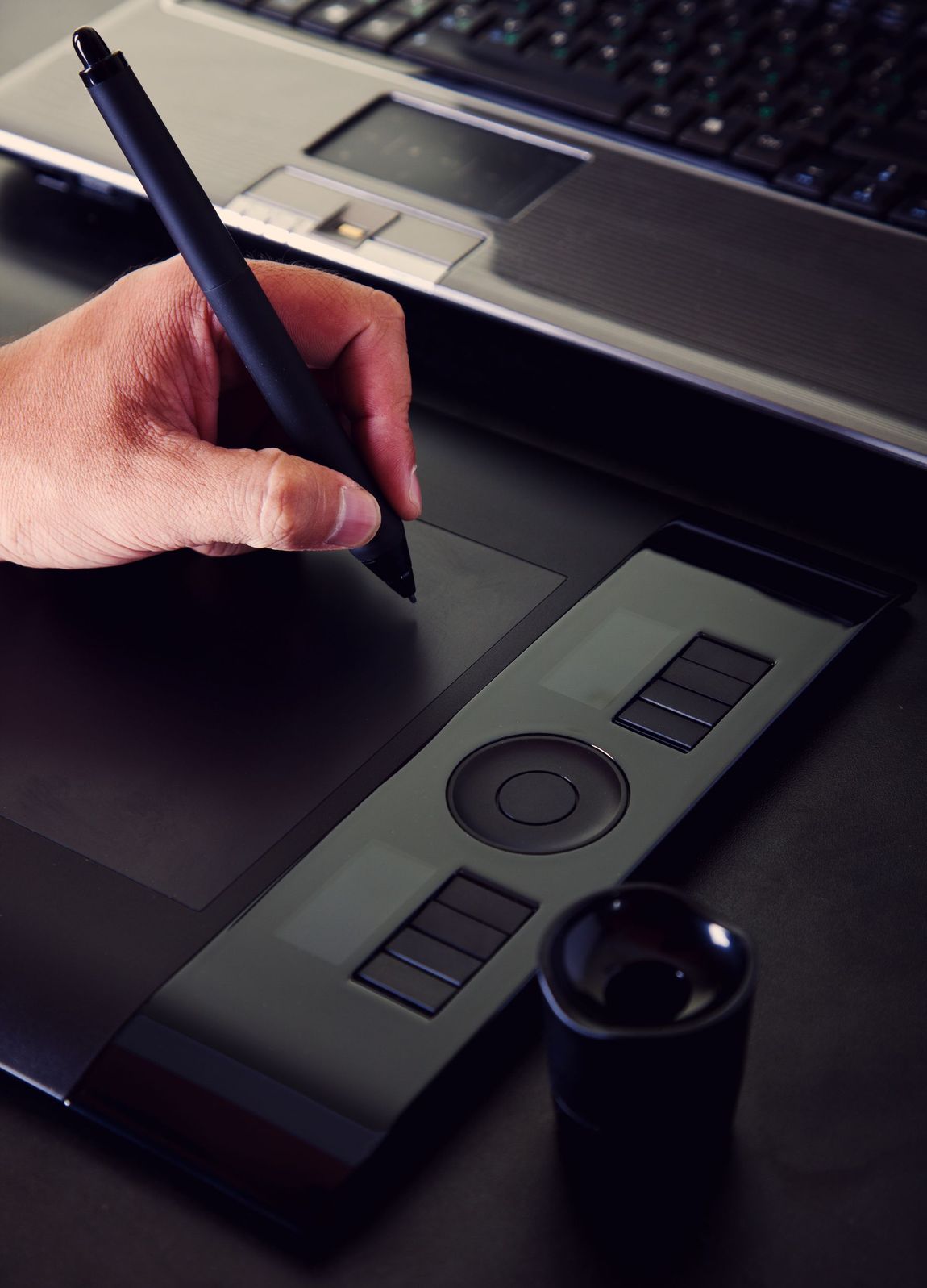 Hand using a stylus on a black graphics tablet next to a laptop.