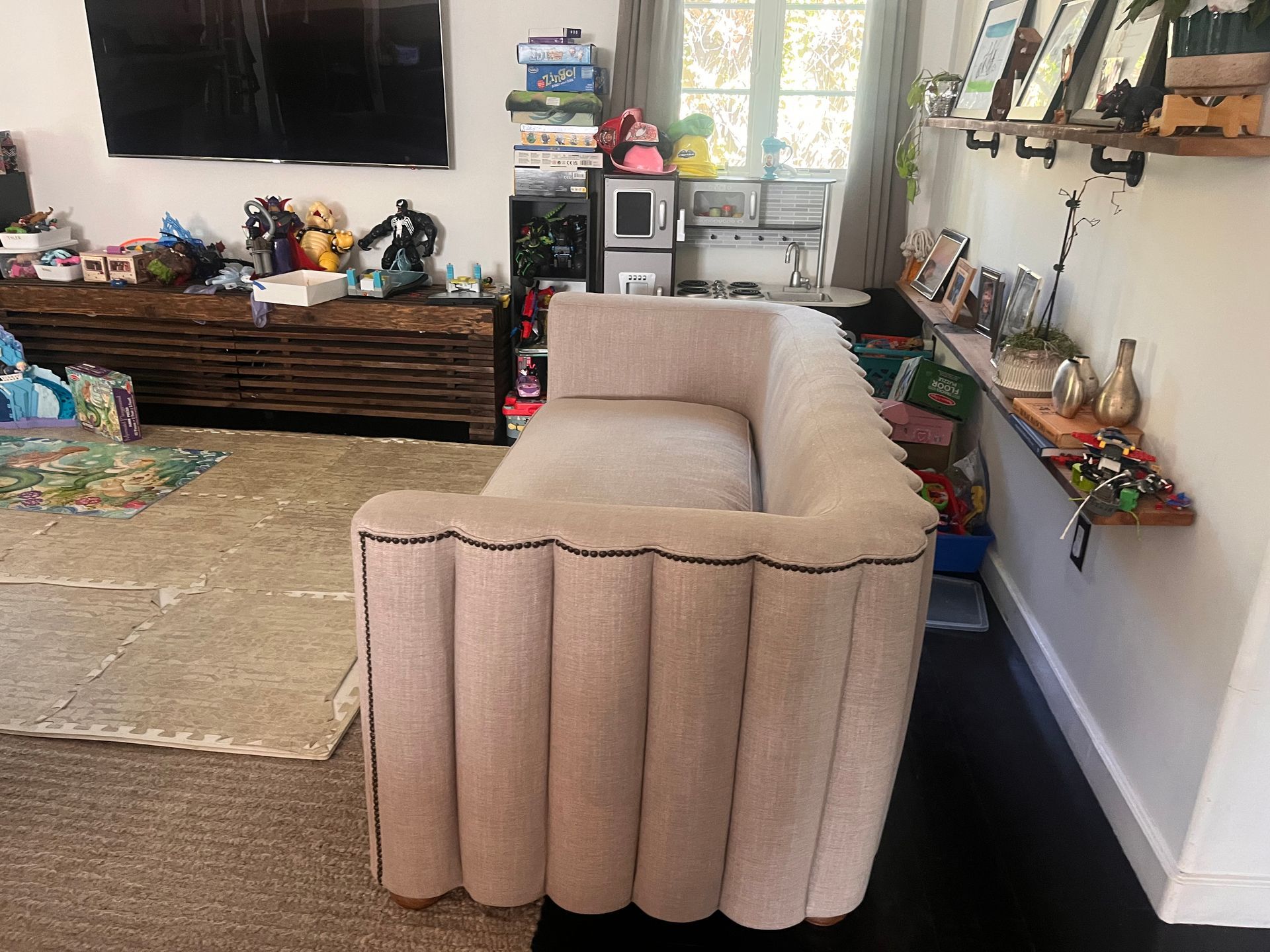 Beige sofa in a cluttered living room with a TV, shelves, and a rug on the floor.