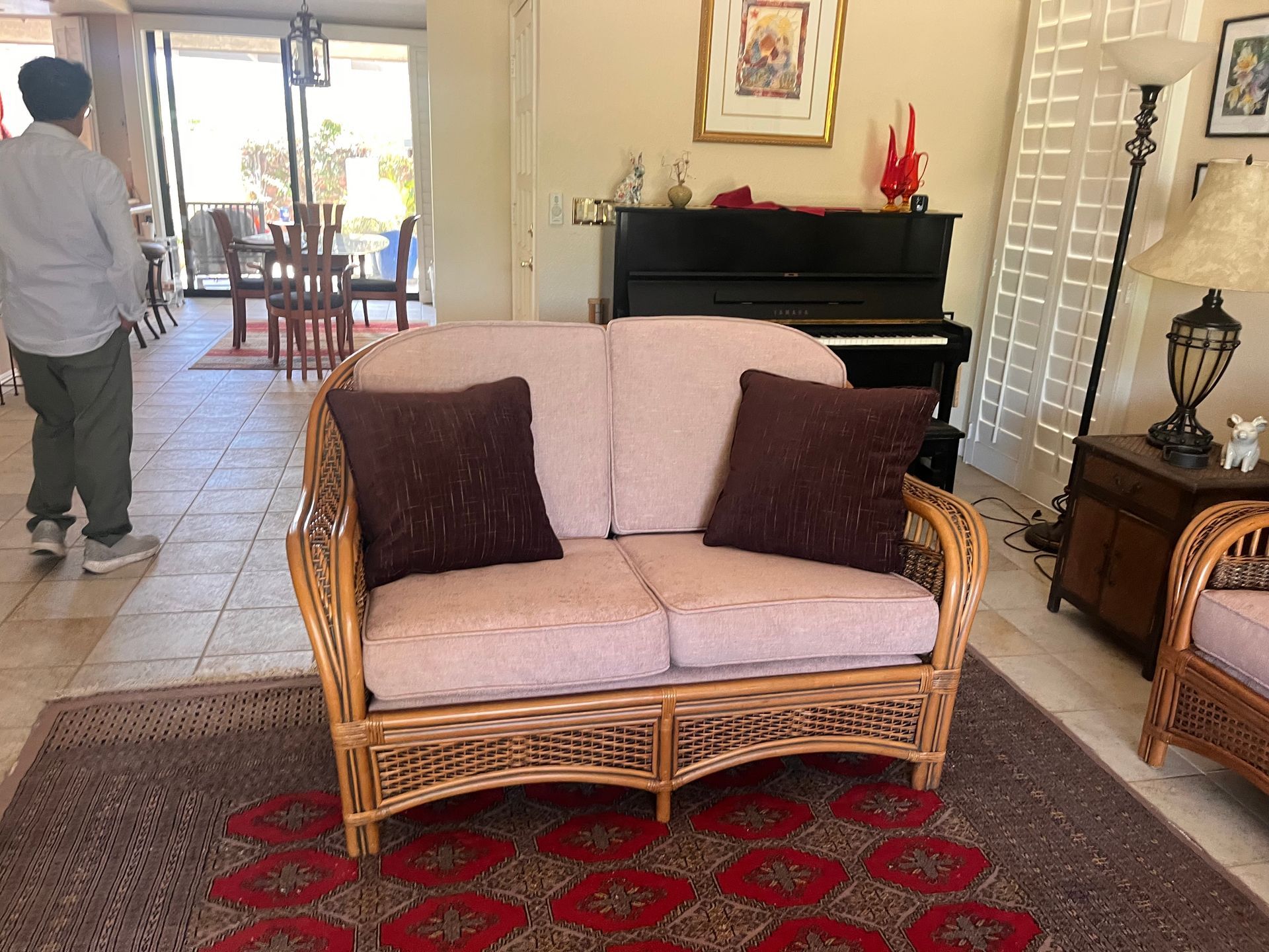 Living room with wicker loveseat, red rug, piano, and a person standing near the doorway