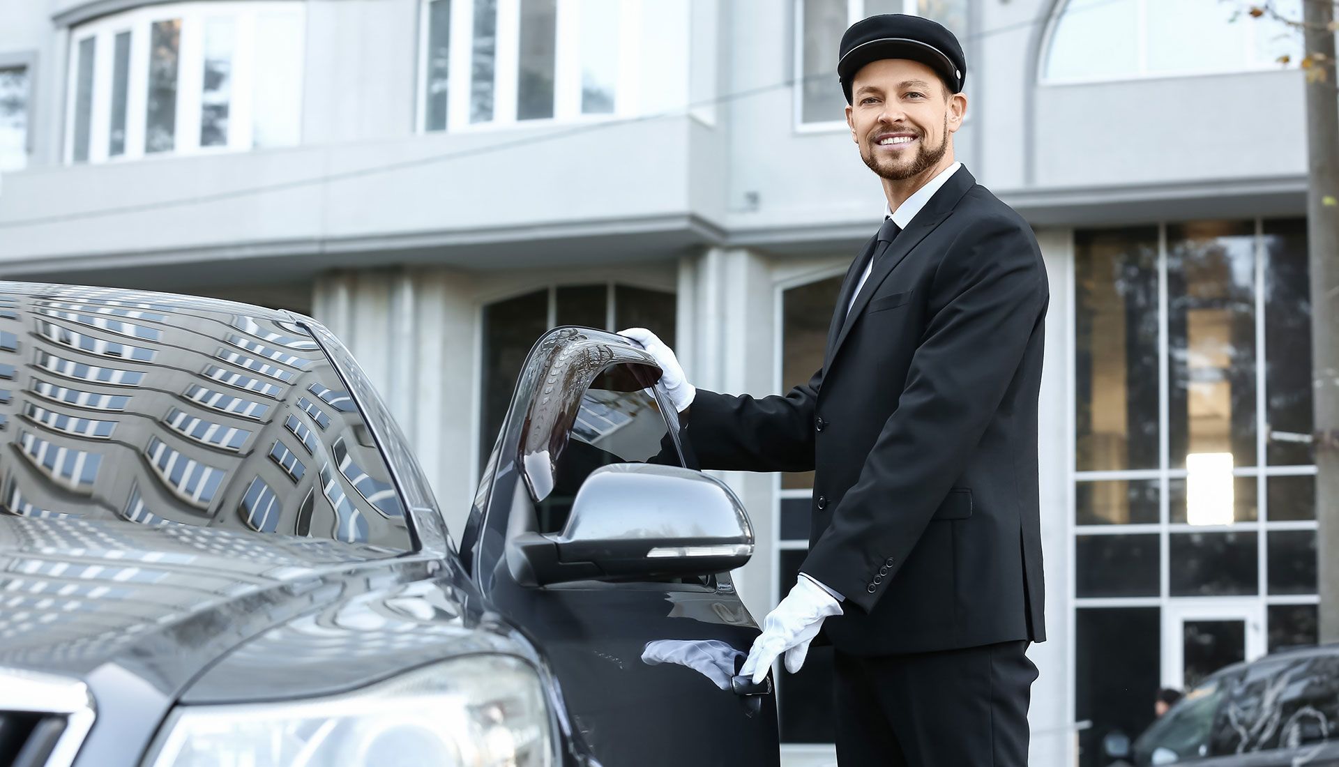 A chauffeur wearing a black uniform and cap polishing a black car's side mirror outside a building.