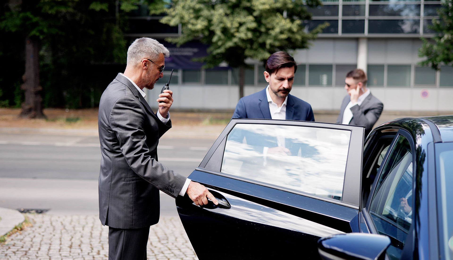 Man in suit opening car door for two other men in suits, outdoors.