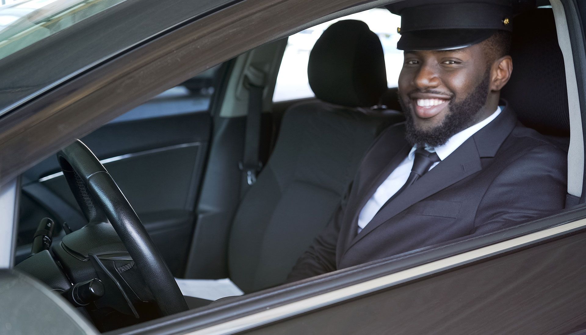 Man in chauffeur uniform smiling in a car, seated behind the steering wheel.