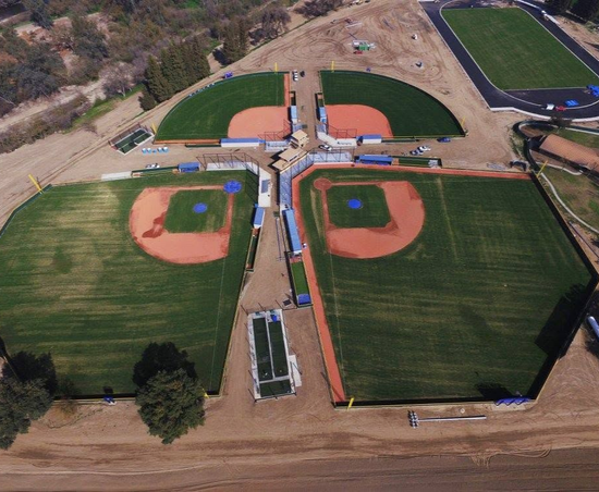 Aerial view of four baseball fields with green grass, red infields, and a central walkway/dugout complex.