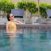 Woman relaxing in a pool, wearing a yellow swimsuit and sunglasses, with lounge chairs and greenery behind her