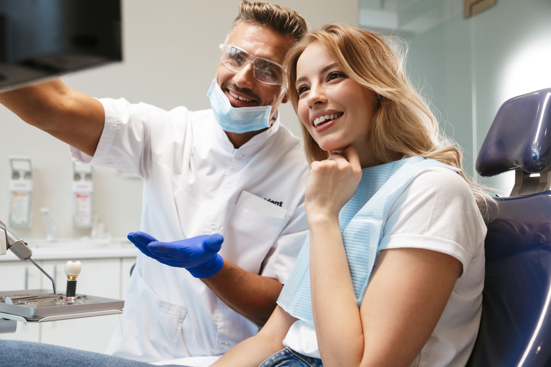 A smiling young woman sits in a medical dentist chair, looking at an x-ray scan, next to a dentist. A smiling young woman sits in a medical dentist chair, looking at an x-ray scan, next to a dentist.