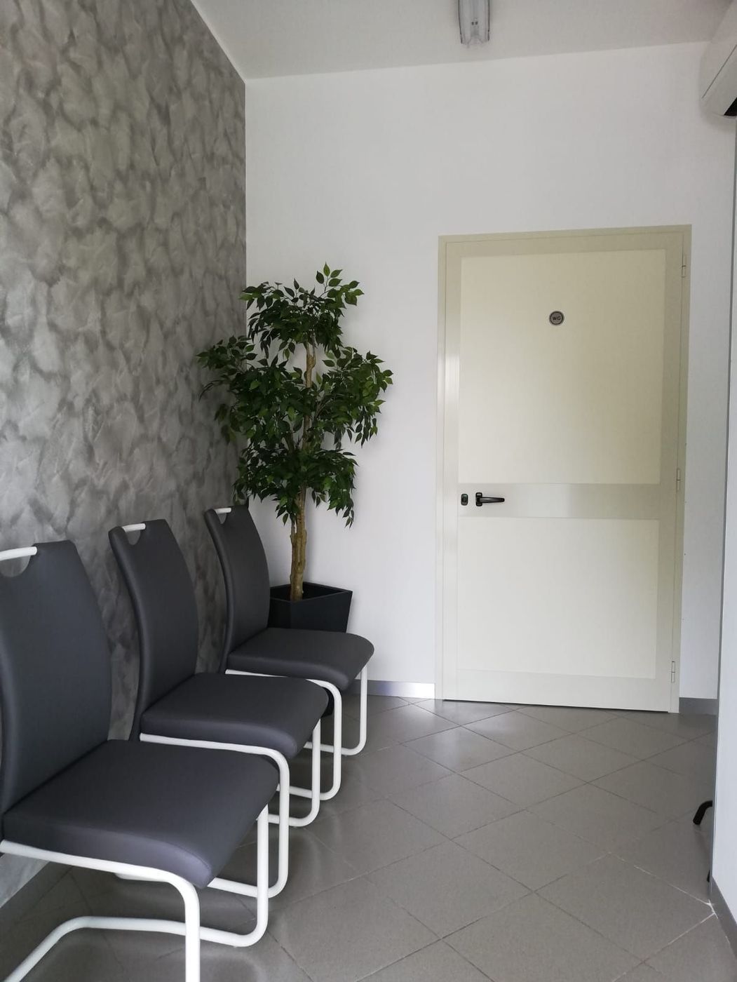 Sala d'attesa con sedie nere, pianta in vaso e porta bianca in un corridoio piastrellato.