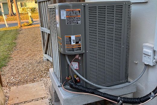 Air conditioning unit on a concrete base next to a white wall, with electrical conduit and a gravel ground.