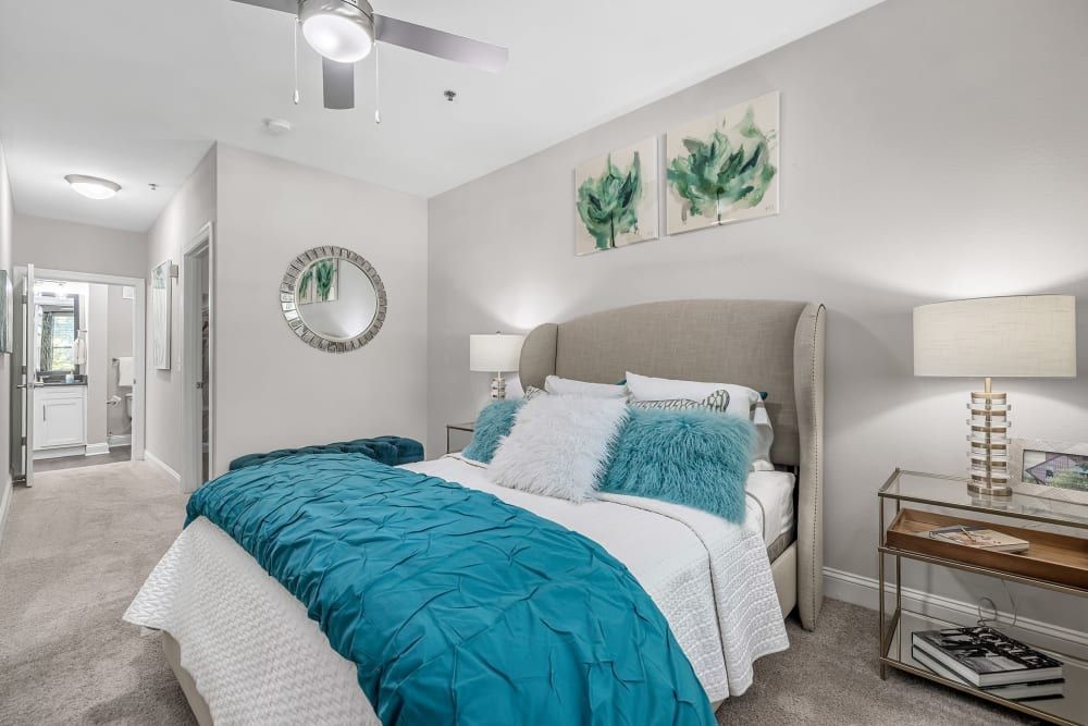 Modern bedroom interior with a neatly made bed covered in a white and turquoise bedding set, complemented by fluffy turquoise pillows. Two framed abstract green art pieces hang above the bed, matching the color theme. A mirrored bedside table houses a contemporary lamp, next to a window with closed white blinds. Across the bed, an open door leads to an attached bathroom, and a stylish circular mirror hangs on the adjacent wall at Marq Perimeter in Atlanta, GA.