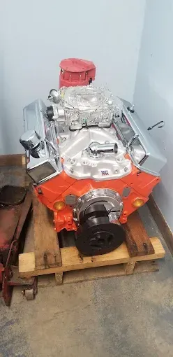 An orange V8 engine with polished chrome valve covers and a silver intake manifold sitting on a wooden pallet.