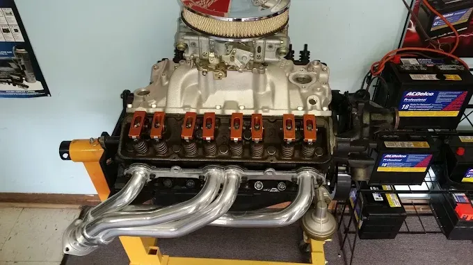 A disassembled V8 engine mounted on a yellow stand, featuring chrome headers, a silver intake, and red rocker arms.