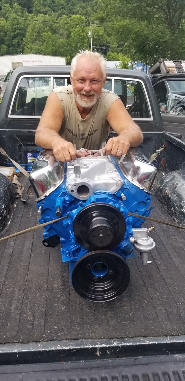 A smiling person with a beard rests their arms on a freshly painted, bright blue V8 engine sitting in a truck bed.