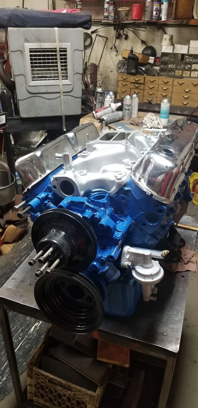 A blue V8 engine with polished chrome valve covers and a silver intake manifold sitting on a metal workbench in a shop.