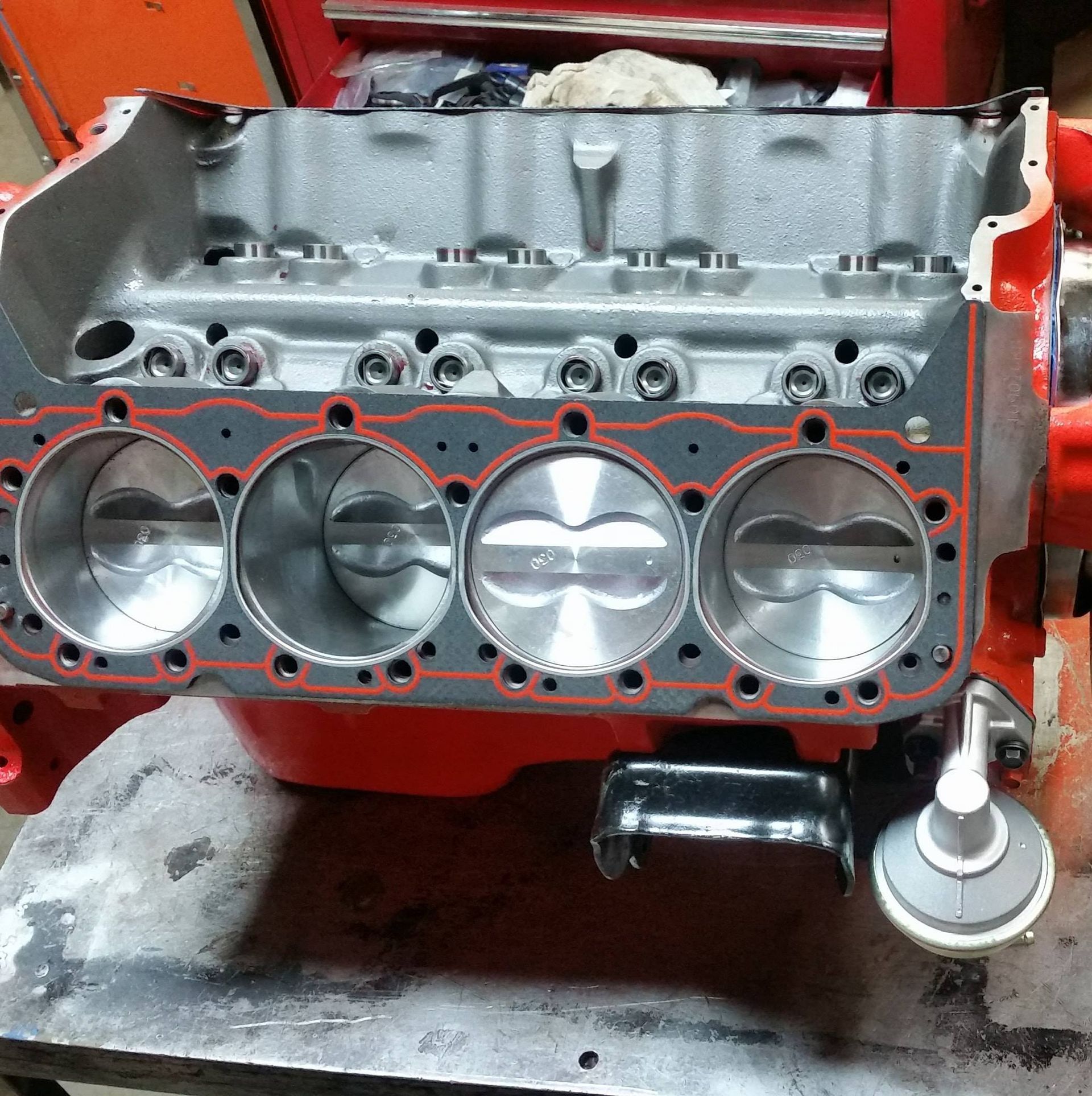 A partially assembled red V8 engine block with a head gasket and pistons visible, sitting on a workbench.