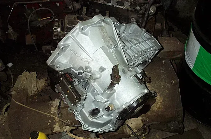 A clean, silver-colored automotive transmission assembly sitting on a cluttered workshop floor.
