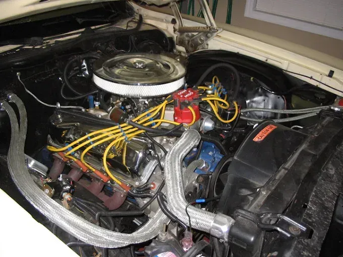 An open hood reveals the engine bay of a classic car, highlighting a round air cleaner, yellow spark plug wires, and hoses.