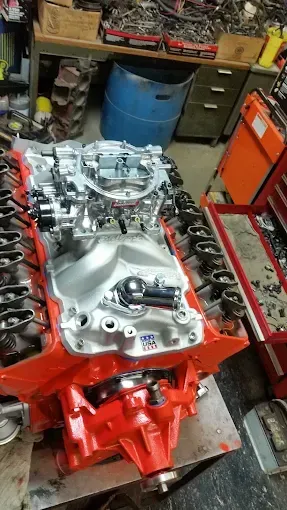 A partially assembled V8 engine block painted bright orange with an aluminum intake manifold and carburetor on top.