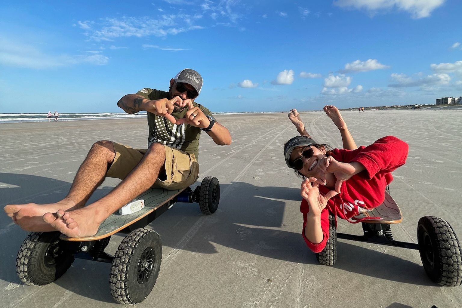 Two people riding off-road skateboards on a beach, one in tan and one in red, under a blue sky