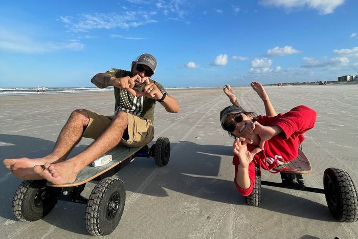 Two people riding off-road skateboards on a beach, one in tan and one in red, under a blue sky