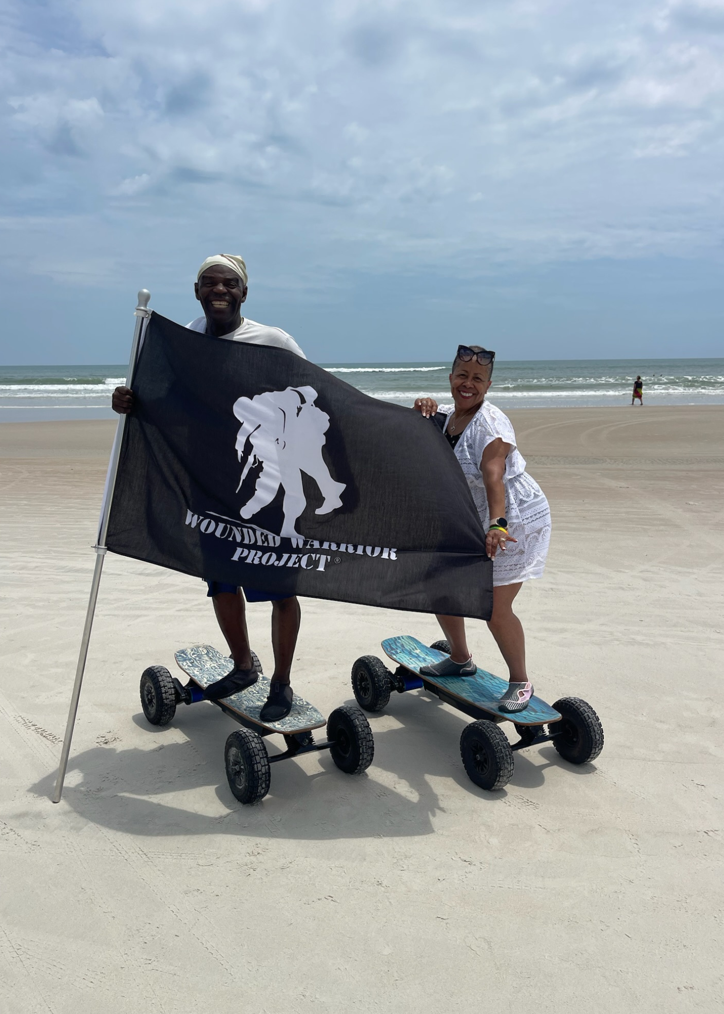 Two people posing on electric scooters at a beach, holding a black flag under a cloudy sky