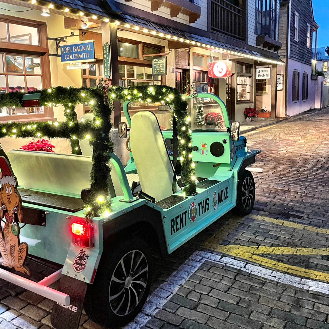 Light-green golf cart decorated with lights, parked on a cobblestone street in St.Augustine, next to shops with lights