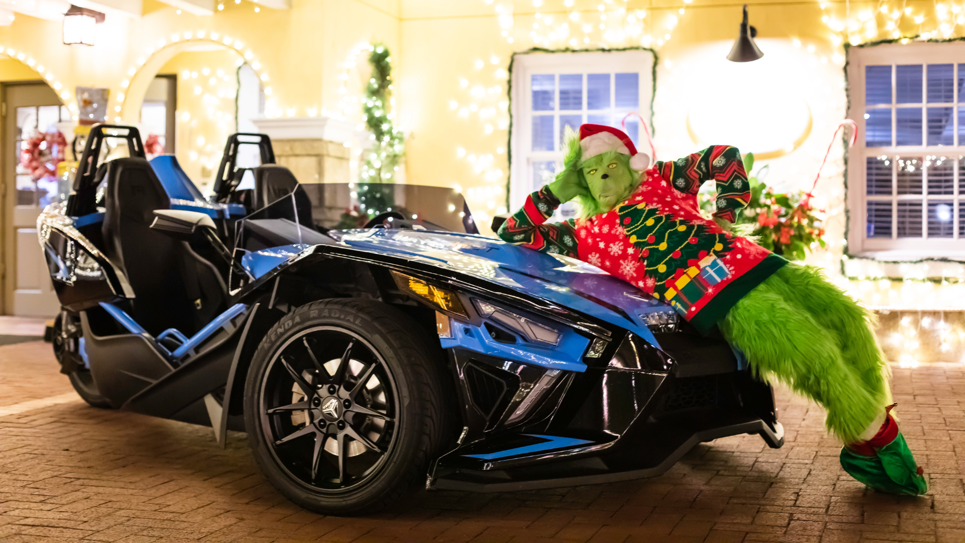 Grinch in Christmas sweater leaning on a blue three-wheeled Slingshot vehicle outdoors in St. Augustine