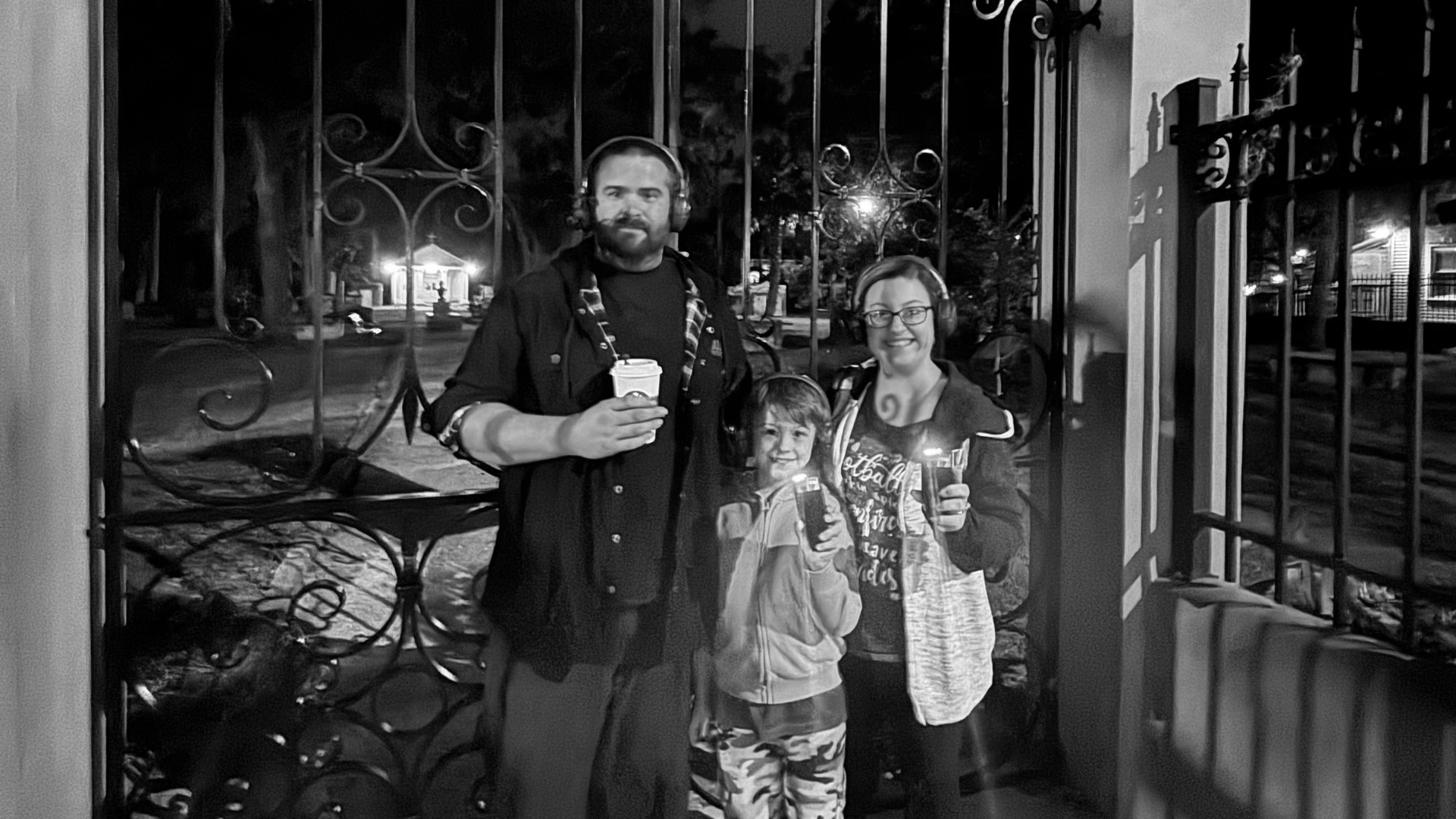 Three people stand at a gate at night, holding drinks, with city lights in the background.