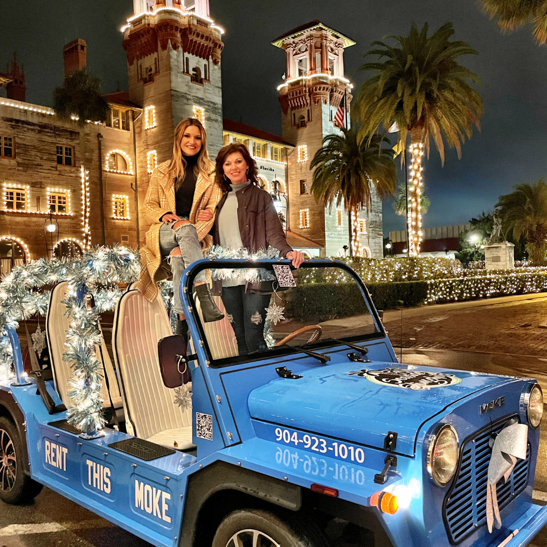 Two women in blue open-top Moke decorated for holidays, in front of a lit up castle in St. Augustine, Florida