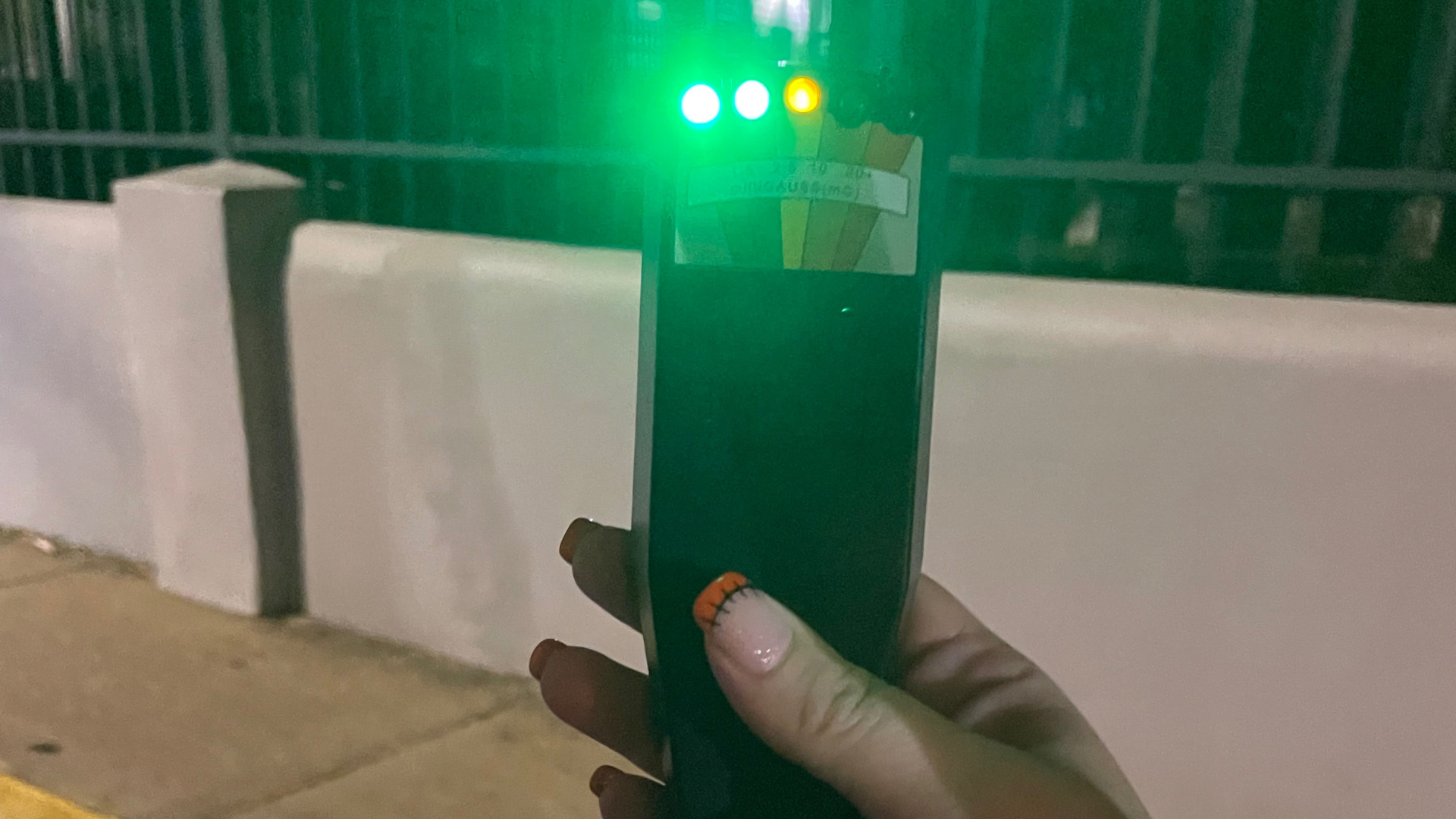 Hand holding a phone at night beside white barriers, with green lights glowing in the background