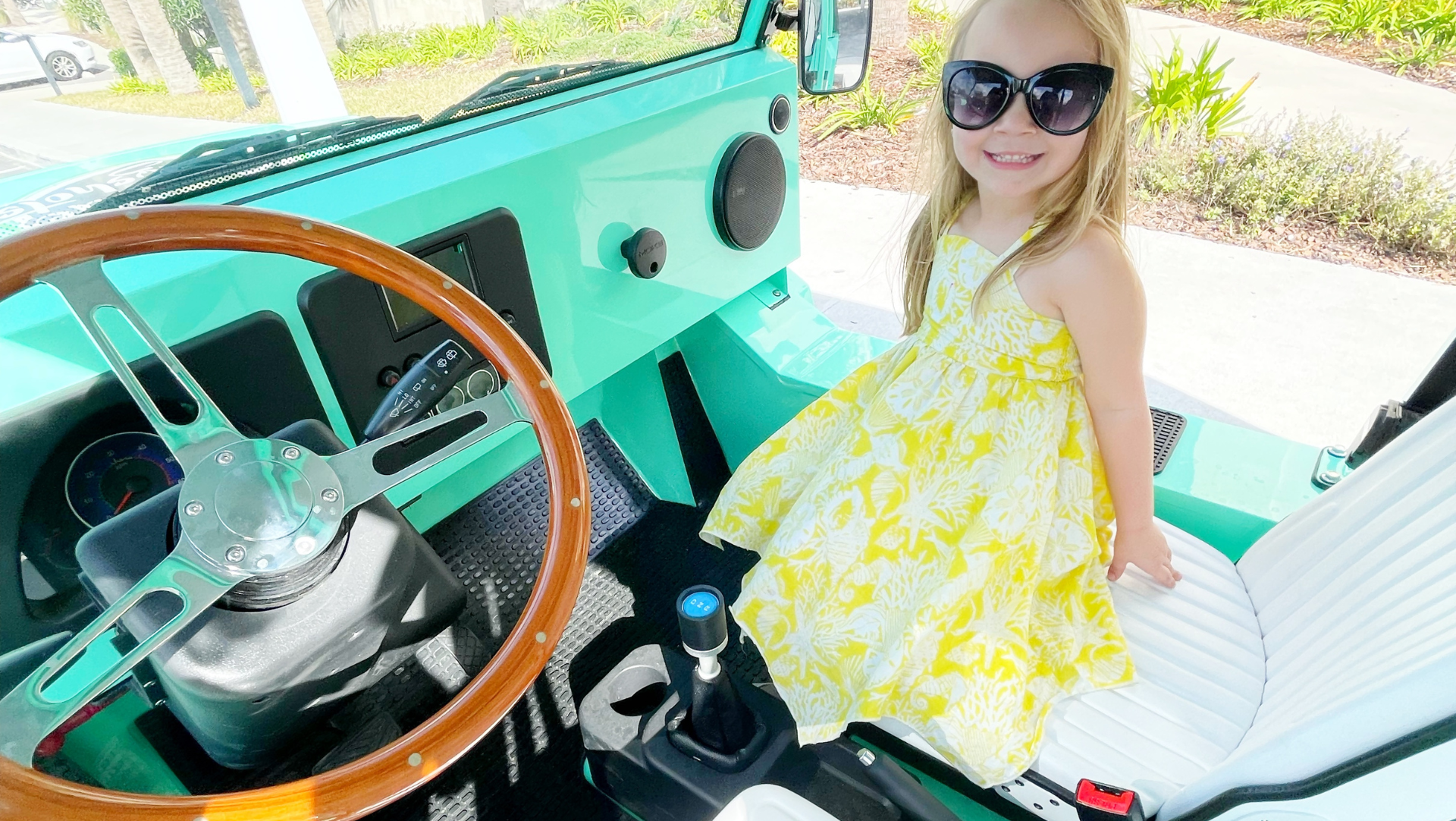 A little girl in a yellow dress is sitting in the passenger seat of a mint electric moke