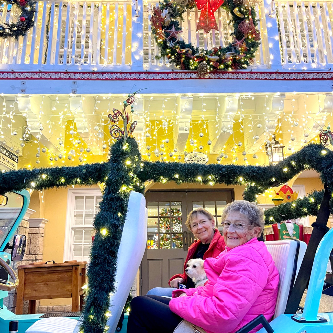 Two women in a teal golf cart in front of a building decorated with Christmas lights and wreaths on a Nights of Lights tour