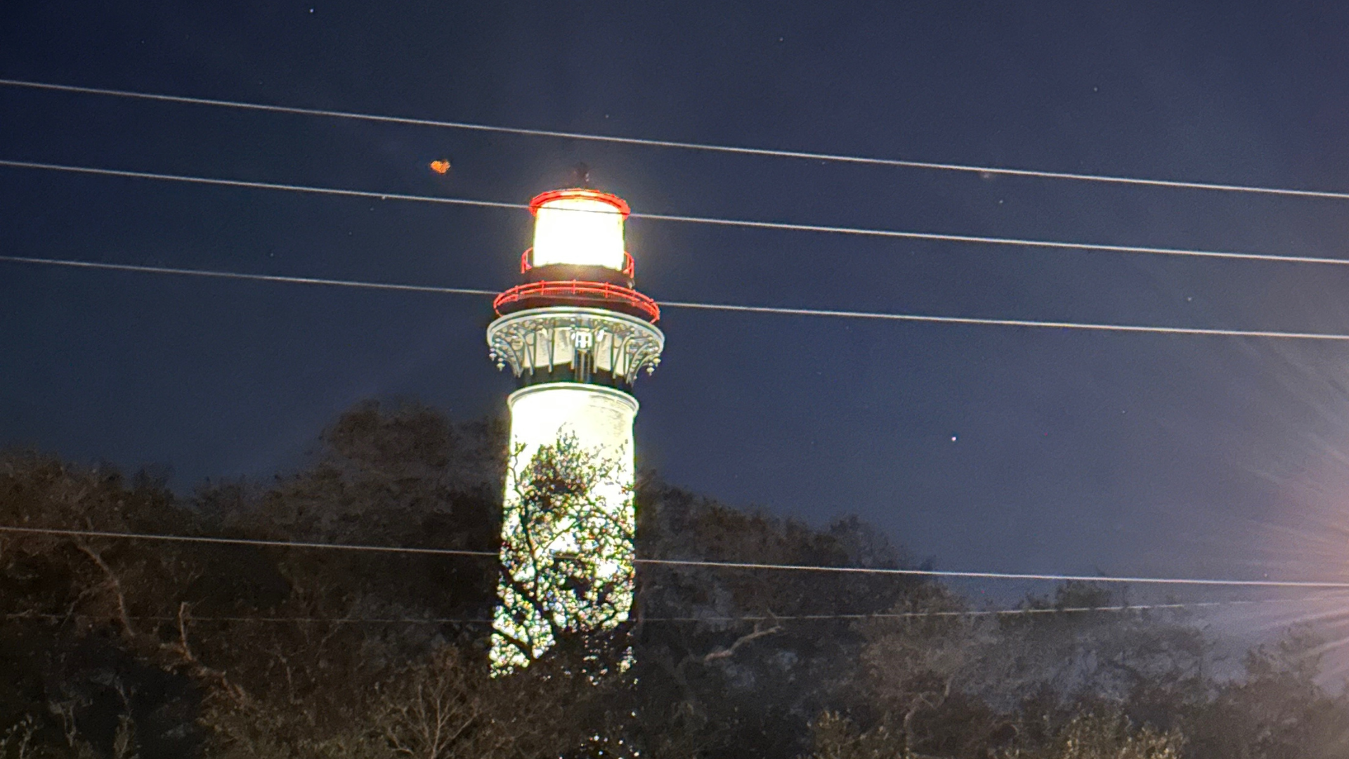 Illuminated lighthouse at night with a red beacon against a dark sky and tree line